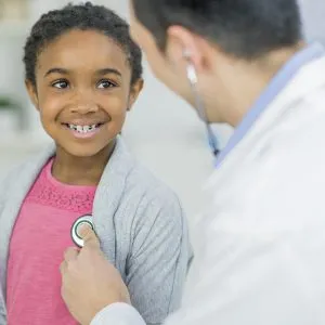 A Caucasian doctor and girl of African descent are indoors in a hospital room. The doctor is wearing medical clothing, and he is checking the girl's heart rate. She is smiling at him.